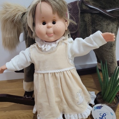 Boneca em vestido bege com coelho bordado, com cavalo de balanço e planta com etiqueta ao lado