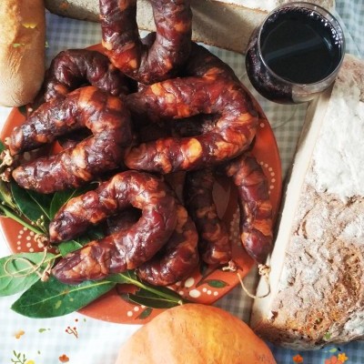 Prato com chouriços, pão rústico, abóbora e copo de vinho tinto sobre toalha xadrez.