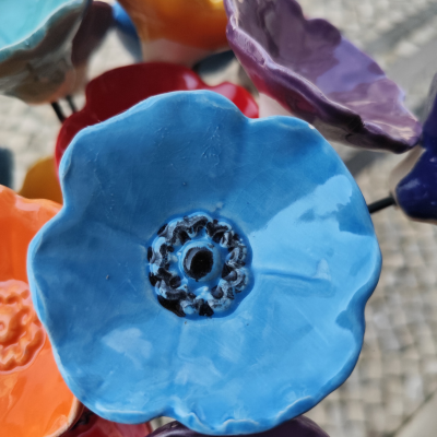 Flor decorativa cerâmica azul com centro preto, em fundo de outras flores coloridas e calçada de pedras