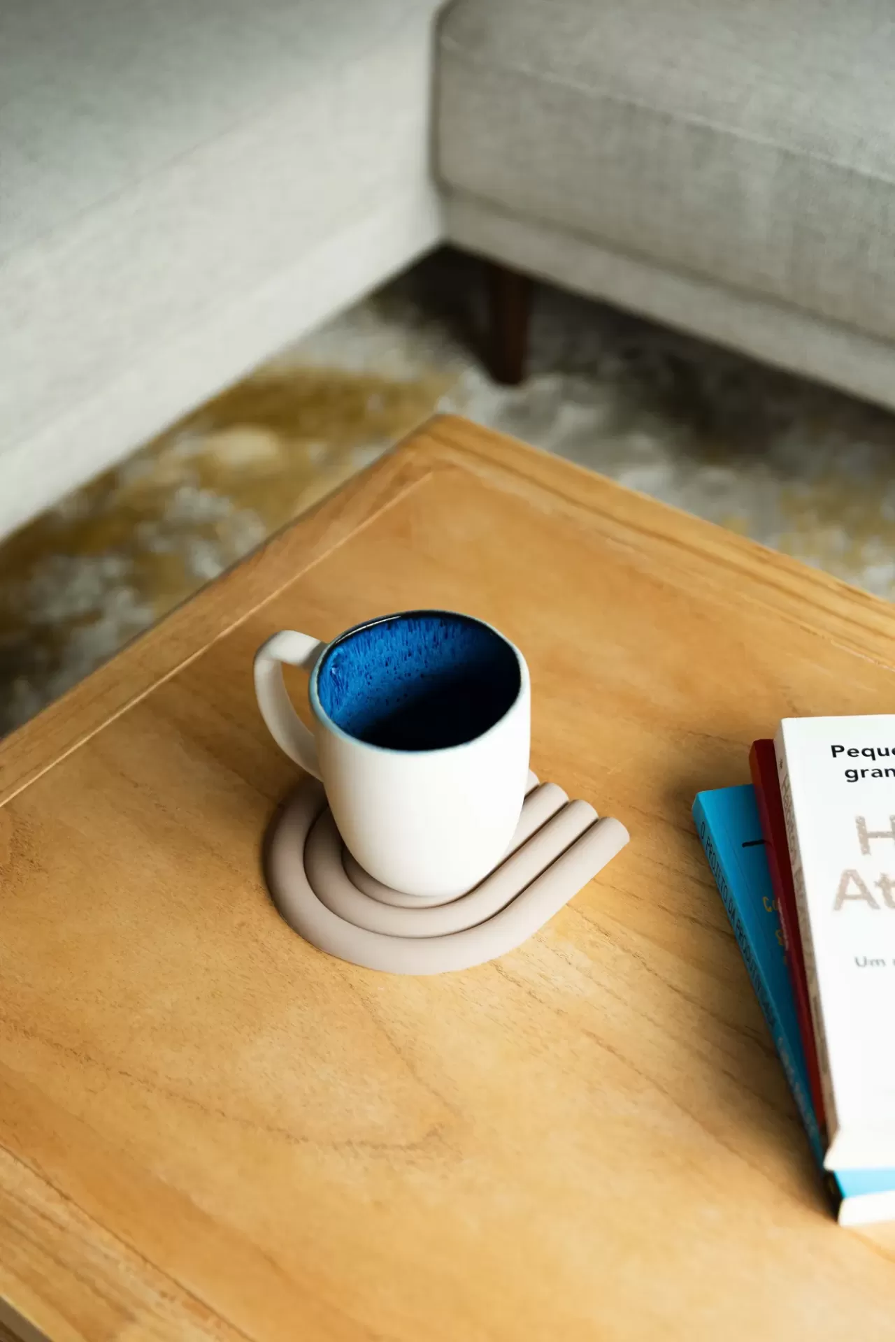 Caneca branca com interior azul sobre suporte de silicone bege numa mesa de madeira clara com livros ao lado.