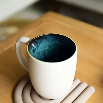 Caneca cerâmica branca com interior azul escuro e base de silicone bege sobre mesa de madeira clara