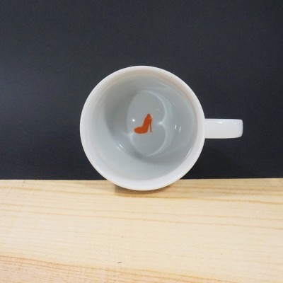 Caneca branca com sapato de salto alto laranja no interior, sobre madeira clara e fundo preto