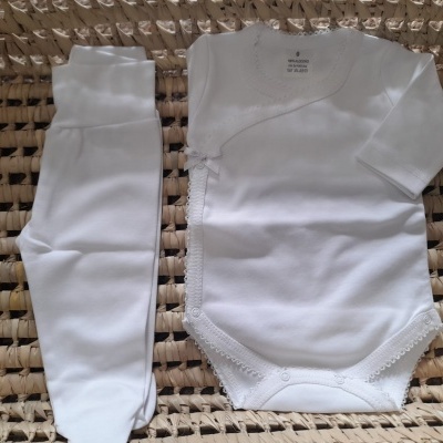 Conjunto de roupa de bebé branco com body e calças sobre suporte de madeira entrelaçada.
