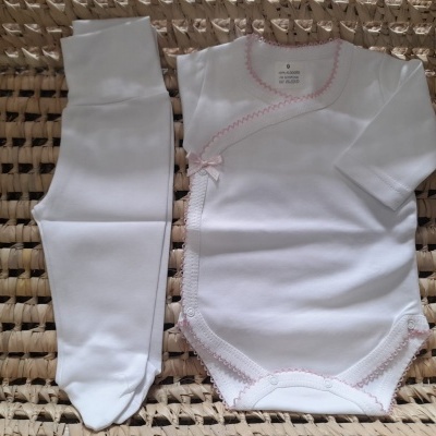 Conjunto branco de roupa para criança com detalhes em rosa sobre superfície tecida bege