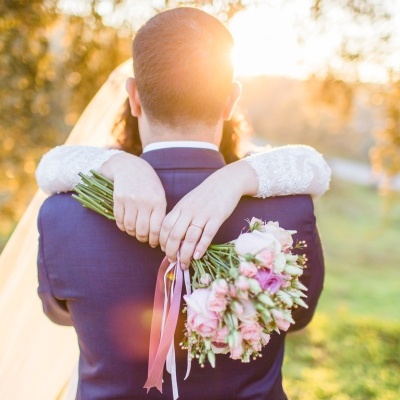 Noivos abraçados ao pôr do sol com ramo de flores cor-de-rosa