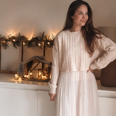 Mulher com camisola de malha bege claro e saia plissada creme, com decoração natalícia iluminada ao fundo