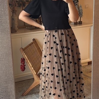 Mulher com roupa preta e saia bege com corações em frente a espelho em sala interior