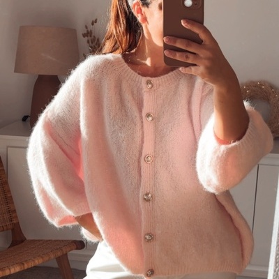 Mulher de cardigã rosa claro com botões prateados e calças brancas a tirar selfie num quarto com cadeira e abajur no fundo.