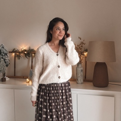 Mulher com cardigan branco e saia comprida castanha com bolinhas em ambiente interior decorado.