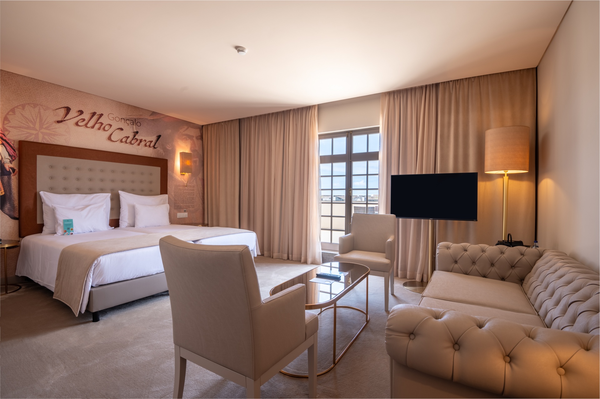 Açores – Nove Ilhas, Um Paraíso Inesquecível Quarto de hotel com camas, poltronas, sofá, mesa de vidro, candeeiro e janela com cortinas