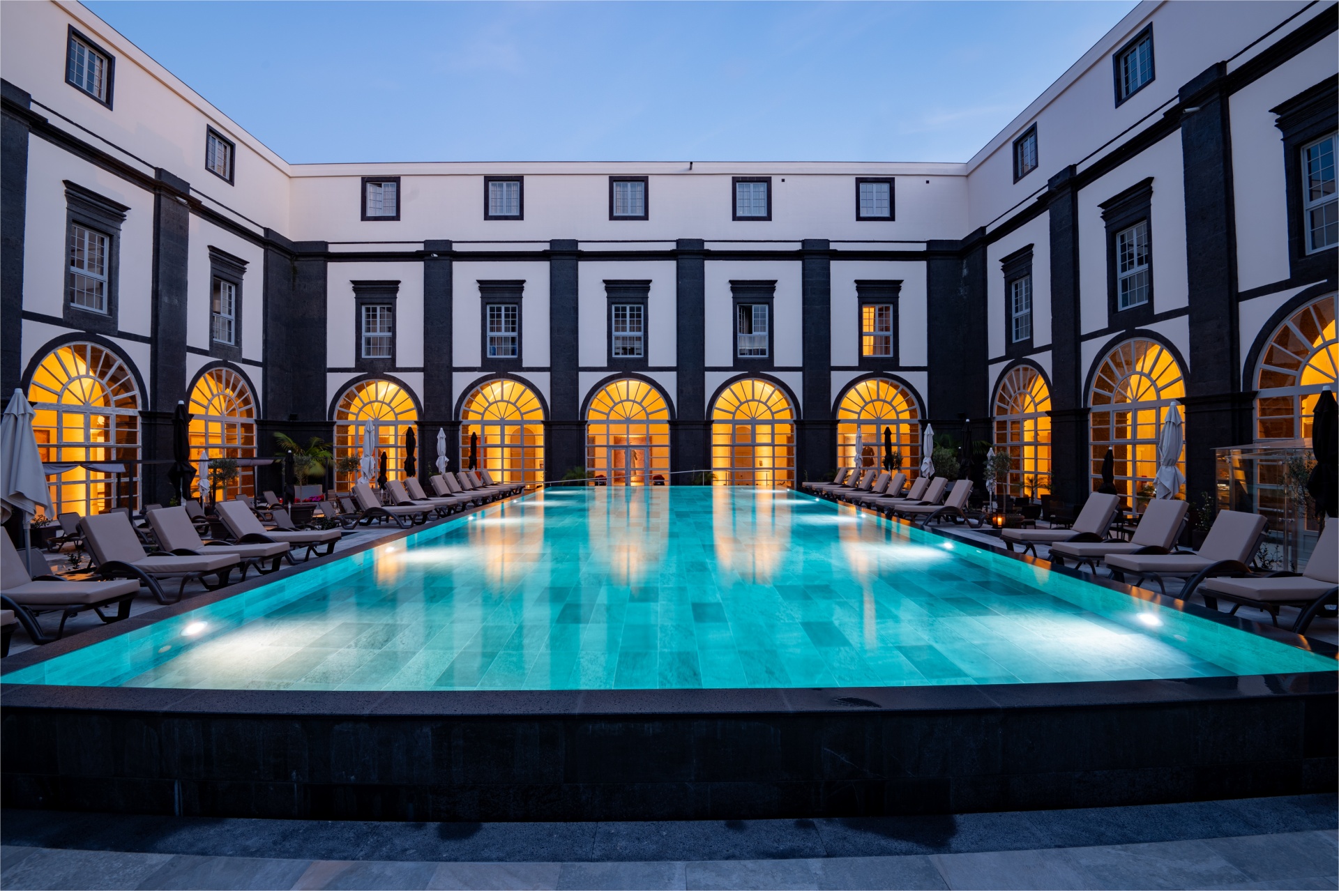 Açores – Nove Ilhas, Um Paraíso Inesquecível Piscina exterior iluminada num pátio de edifício com janelas arqueadas e espreguiçadeiras