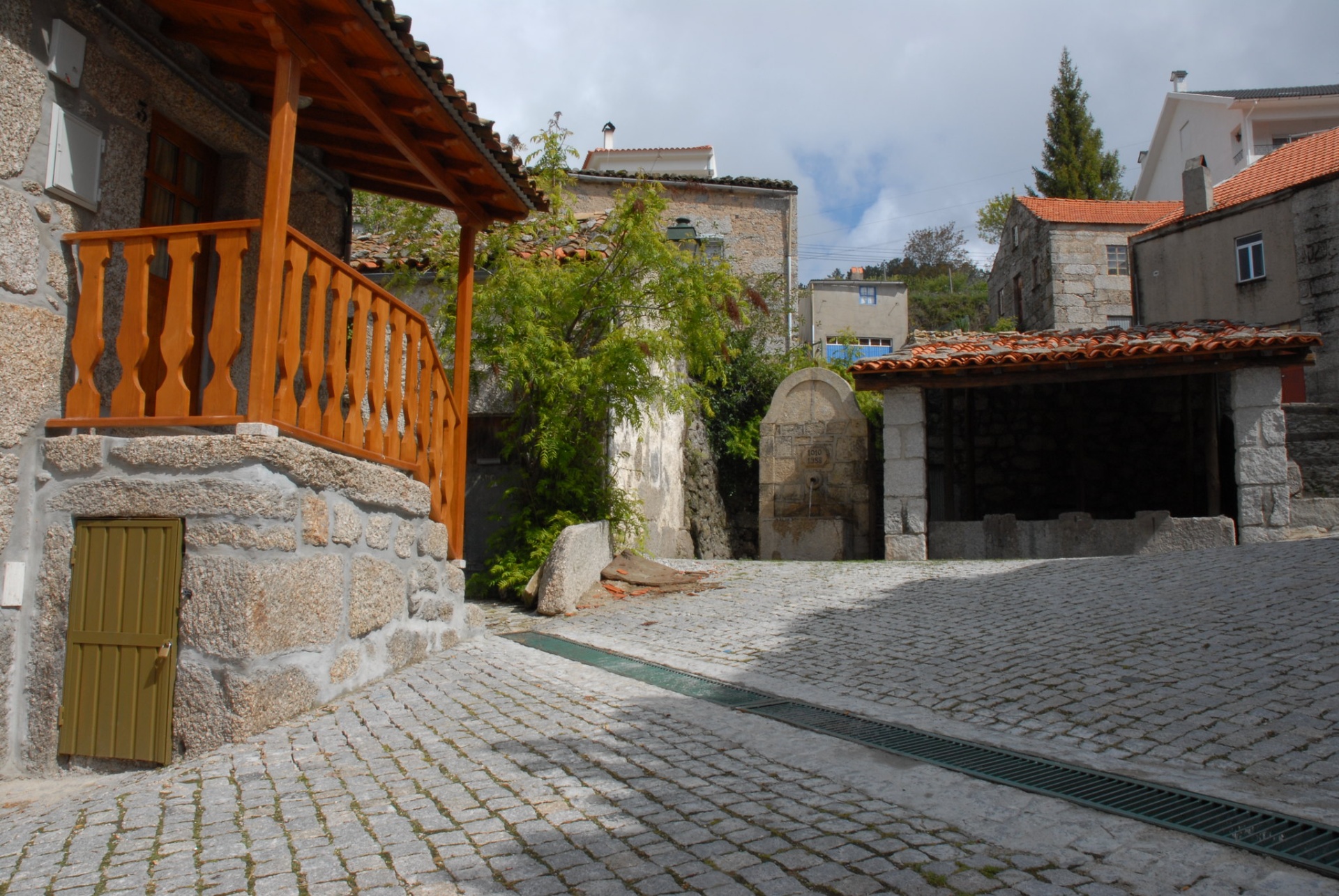 Serra da Estrela – Férias de Natureza e Tradição no Coração de Portugal Área pavimentada com casa de pedra e varanda de madeira, fonte de pedra e edifício com telhado de telhas vermelhas