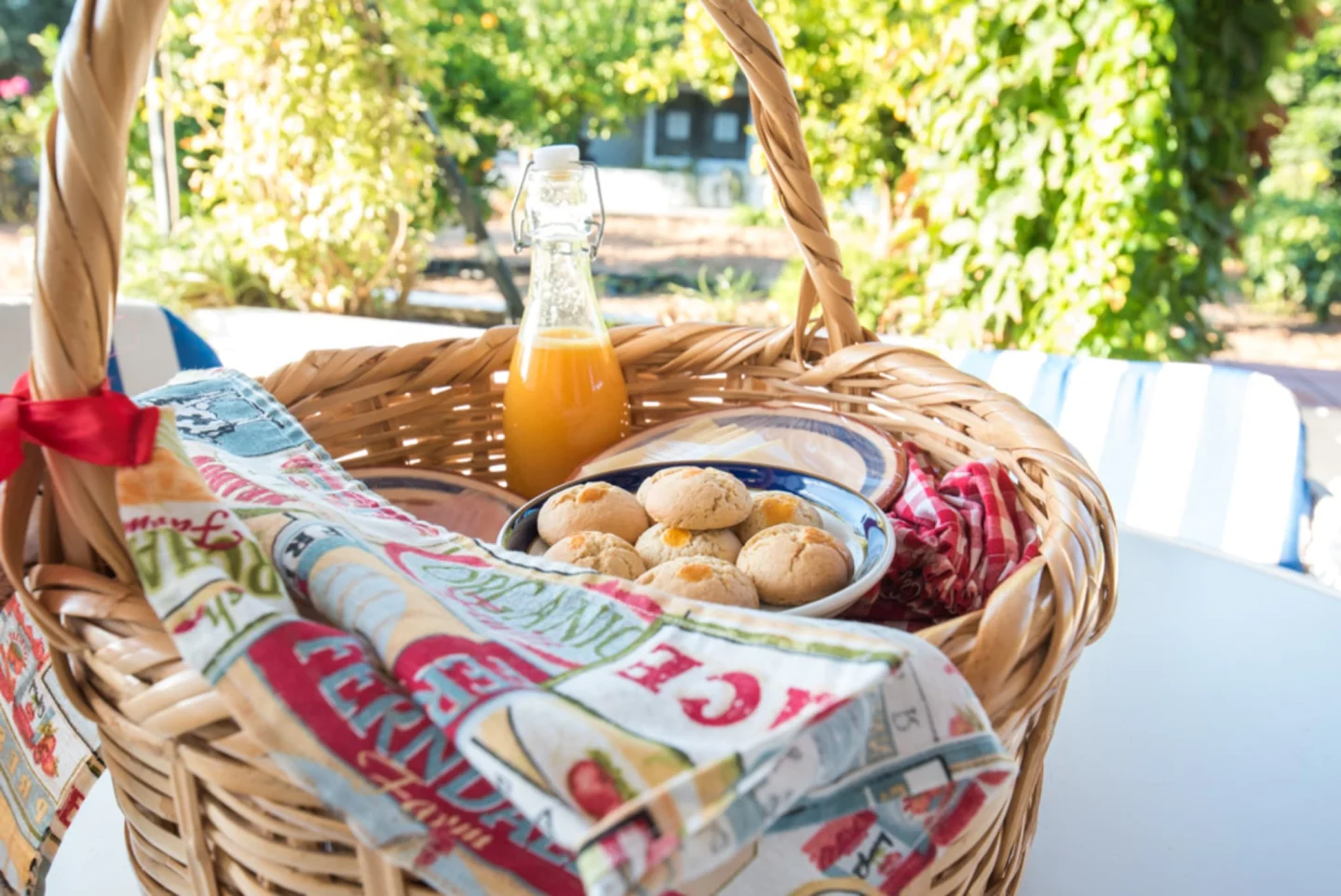 Viagem à medida Cesto de verga com bolachas, frasco de sumo e panos coloridos numa mesa branca ao ar livre