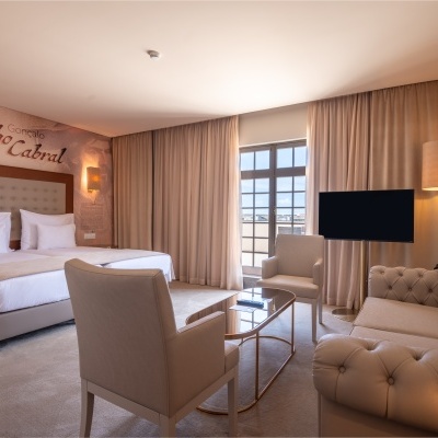 Quarto de hotel com camas, poltronas, sofá, mesa de vidro, candeeiro e janela com cortinas