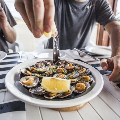 Prato com marisco em conchas aberto a ser temperado com limão numa mesa com toalha riscada azul e branco.