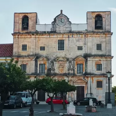 Edifício antigo em pedra com relógio 1924 e sinos em torres, carros estacionados e árvores na frente