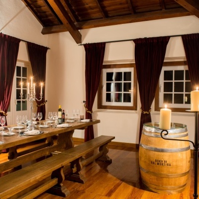 Sala de jantar rústica com mesa de madeira, bancos, velas acesas, cortinas vermelhas e barril de vinho