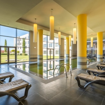 Piscina interior com janelas grandes, colunas amarelas, espreguiçadeiras e luzes pendentes.