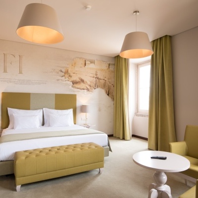 Quarto de hotel com cama de casal, cadeiras e mesa em tons verdes e amarelos e parede decorativa com texto SAFI