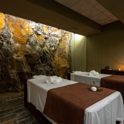 Camas de massagem com lençóis brancos e mantas castanhas em quarto com parede de pedra e iluminação suave.