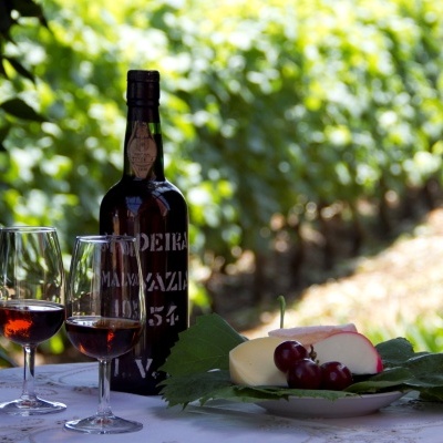 Vinho Madeira em garrafa com duas taças e prato com queijos e uvas numa mesa ao ar livre