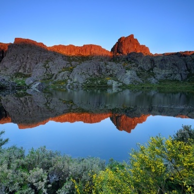 Lago com montanhas rochosas refletidas e vegetação colorida em primeiro plano