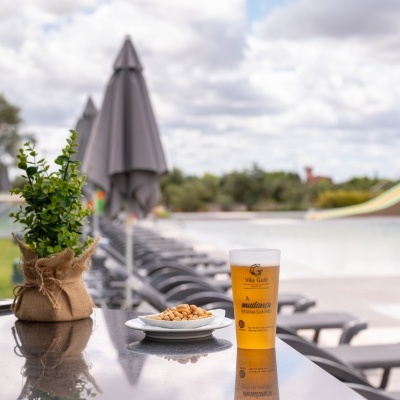 Copo plástico com cerveja e amendoins sobre mesa preta junto a piscina com guarda-sóis cinzentos