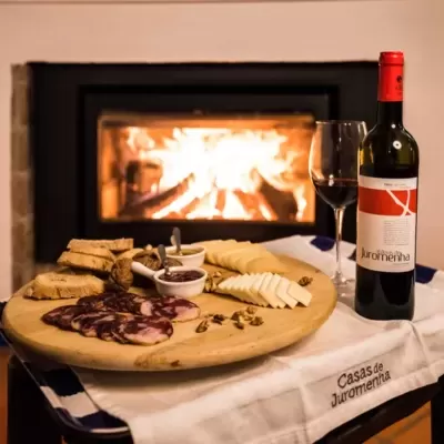 Mesa com vinho tinto, tábua de queijos e enchidos, pão e nozes, diante de lareira acesa