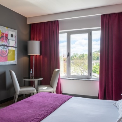 Quarto com cama, cadeiras, mesa, lâmpadas, cortinas roxas e janela com vista