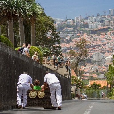 Carrinho de verga artesanal a ser empurrado por duas pessoas em uniforme branco numa rua com vista para cidade e mar