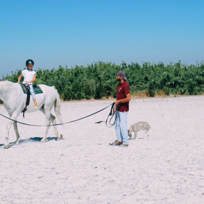 Criança em cavalo branco com guia e homem segurando rédeas num campo arenoso