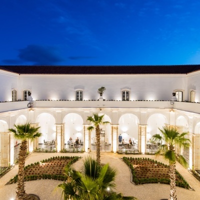 Vista noturna de pátio com paredes brancas, arcos e palmeiras