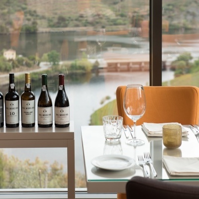 Garrafas de vinho Val Moreira numa mesa branca junto a mesa de jantar com cadeira laranja e vista exterior.