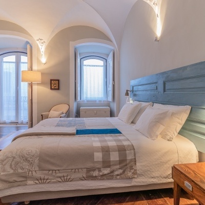 Quarto com cama, cabeceira azul, roupa de cama bege com padrões, janelas com cortinas brancas, mesa de madeira e poltrona