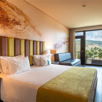 Quarto de hotel com cama, cadeiras verdes, sofá preto, candeeiro, cortinas e vista para colinas