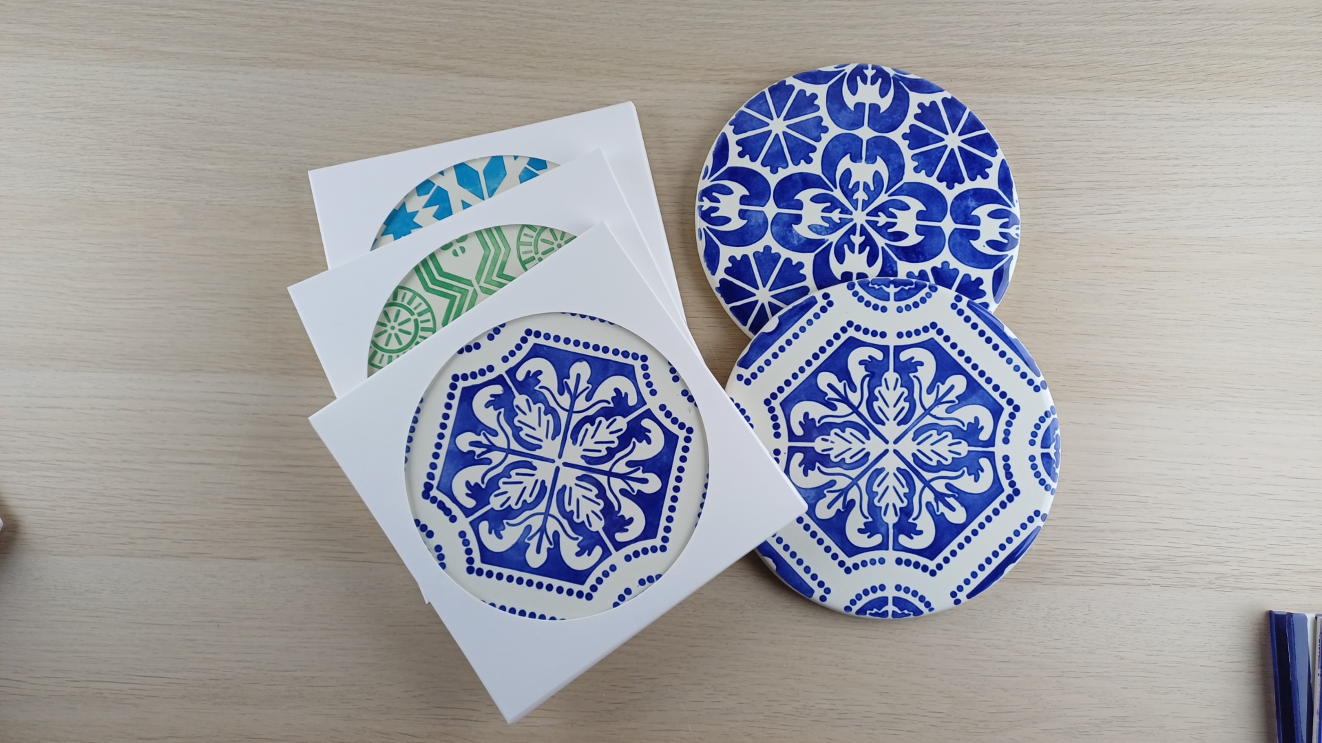 Round ceramic coasters with blue and green patterns, some in white packaging on a light wood surface.