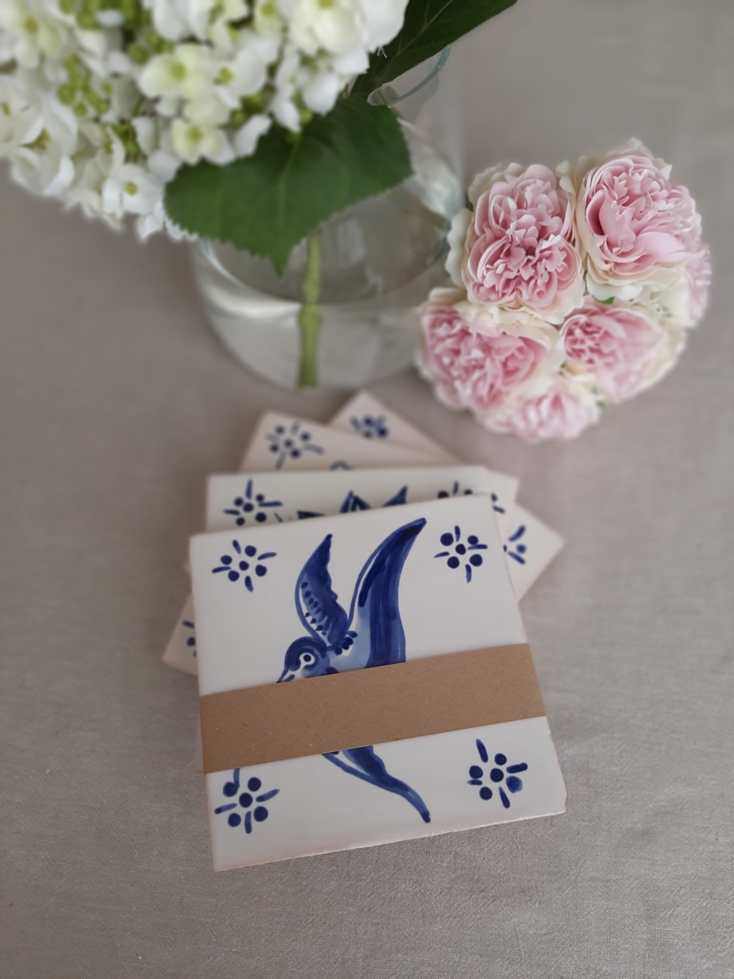 Ceramic coasters with blue bird designs alongside flowers in a vase and pink artificial blossoms.