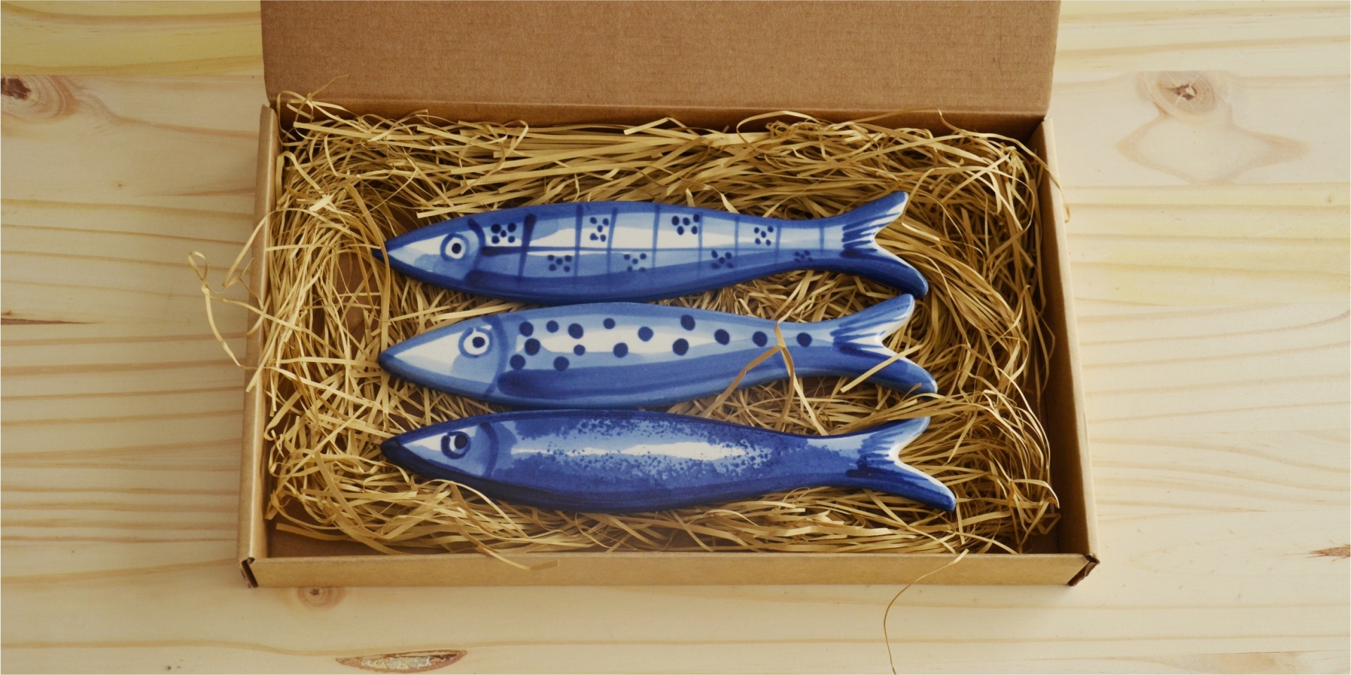 Three blue ceramic fish-shaped items with various patterns in a cardboard box filled with straw.
