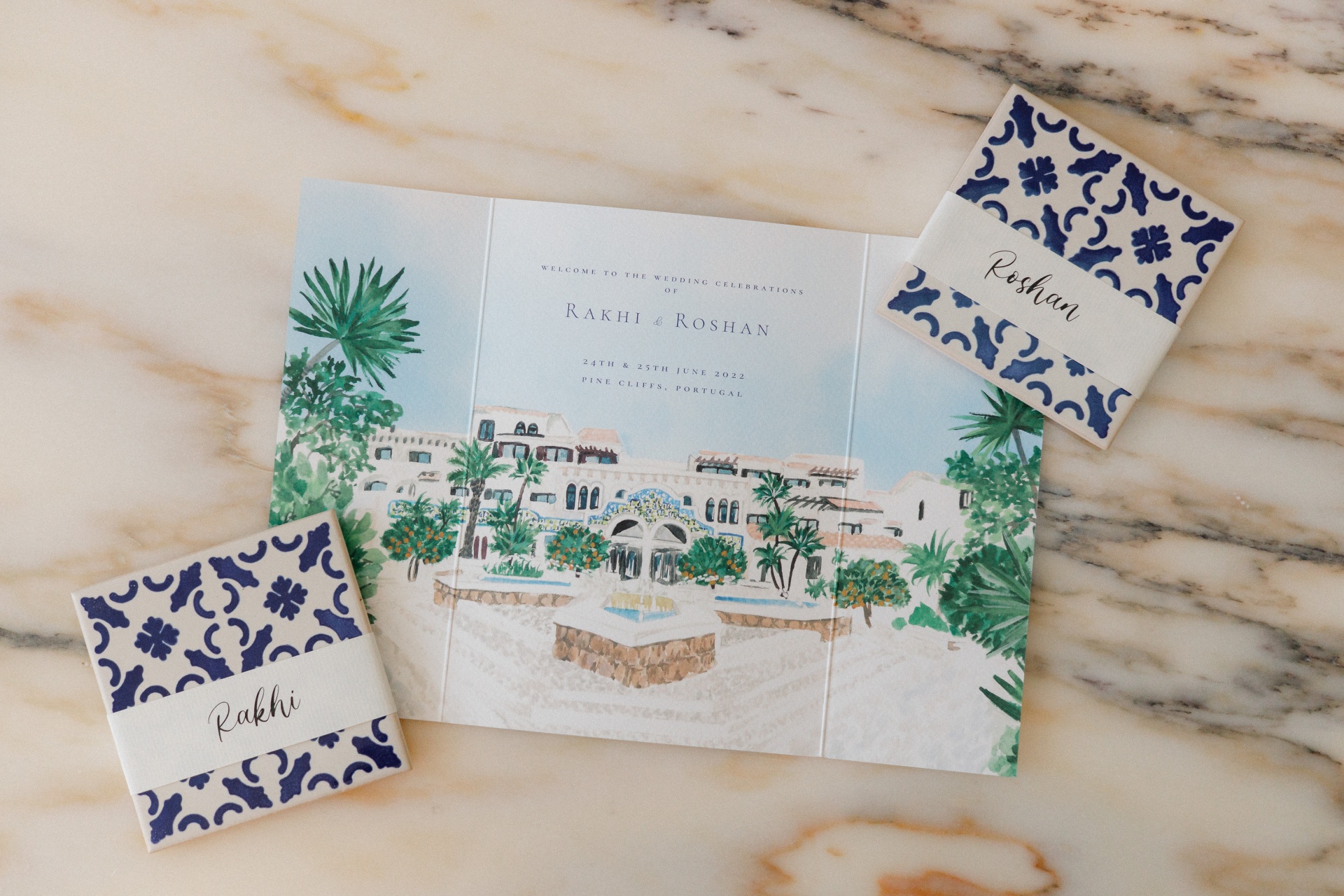 Wedding invitation card with building illustration and two blue patterned name cards labeled Rakhi and Roshan