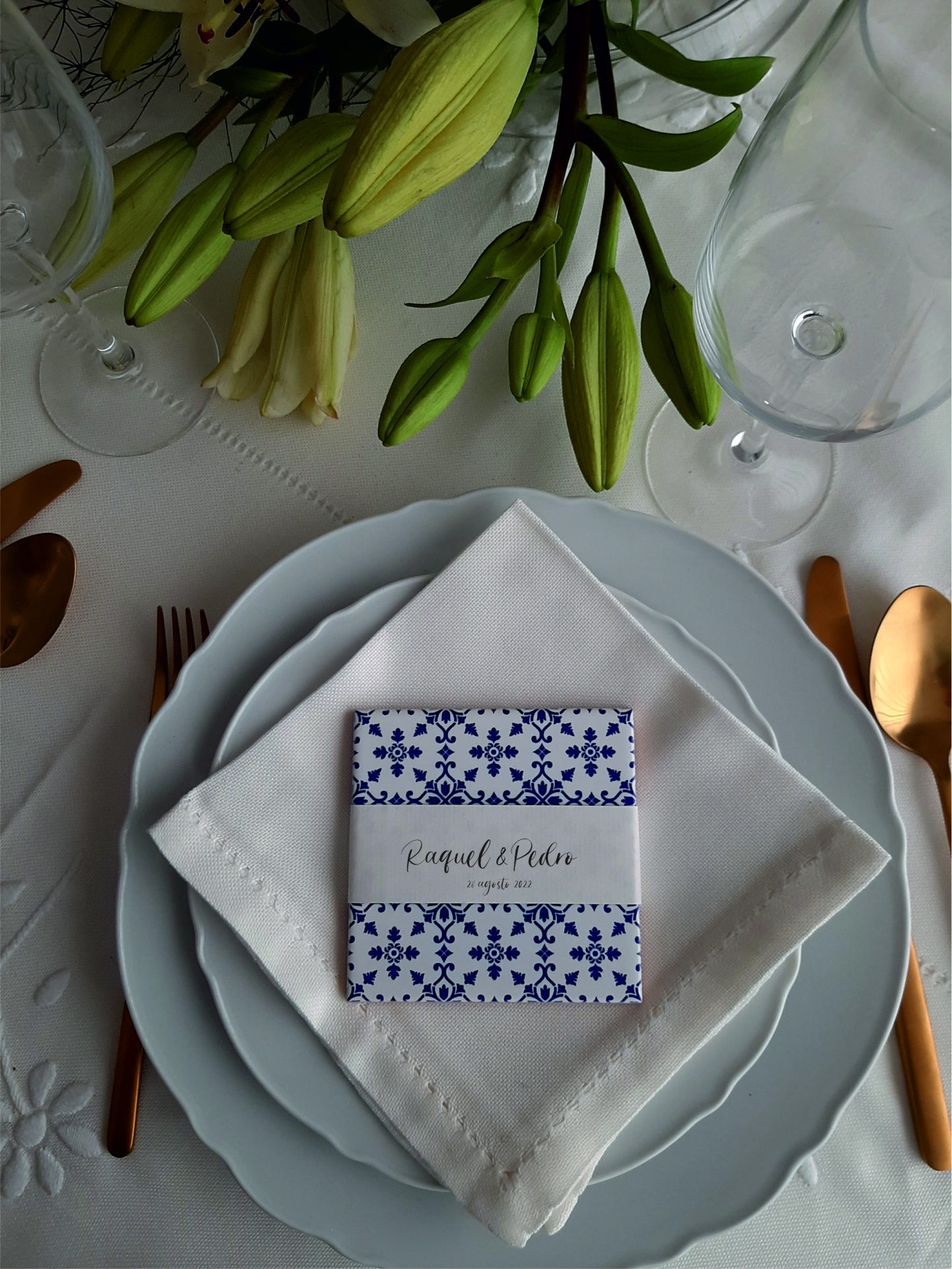 Elegant white table setting with plates, napkin, floral place card, gold cutlery, wine glasses, and lily buds