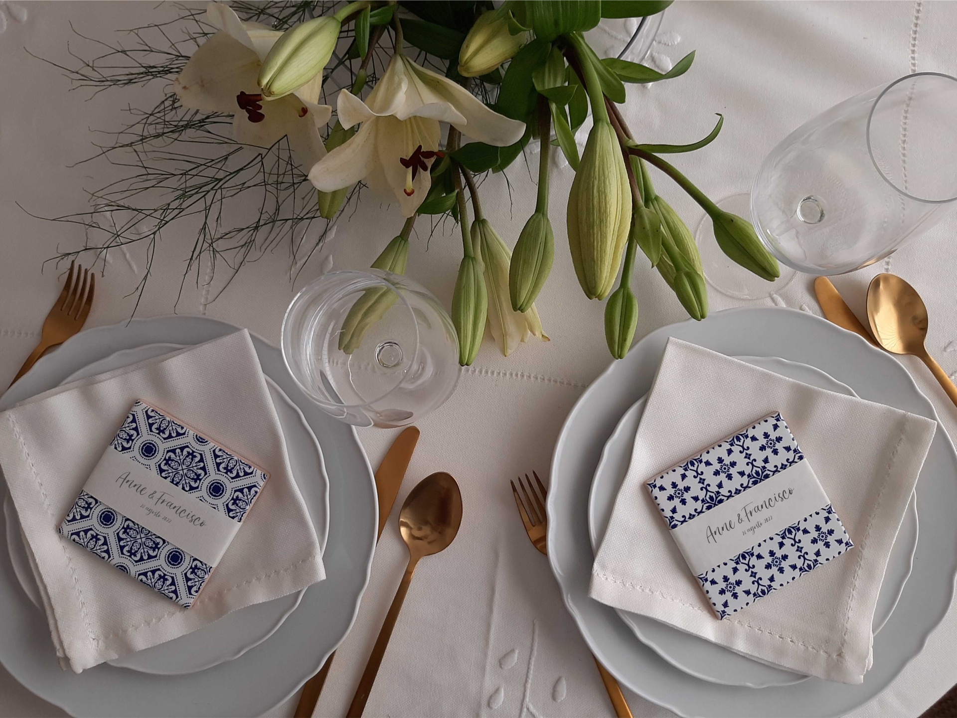 Elegant table setting with white plates, napkins, blue patterned cards, gold cutlery, wine glasses, and lily centerpiece