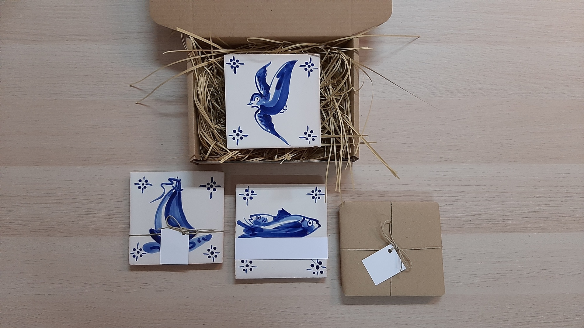 Ceramic tiles with blue bird, fish, sailboat designs and a wrapped blank tile in brown paper.