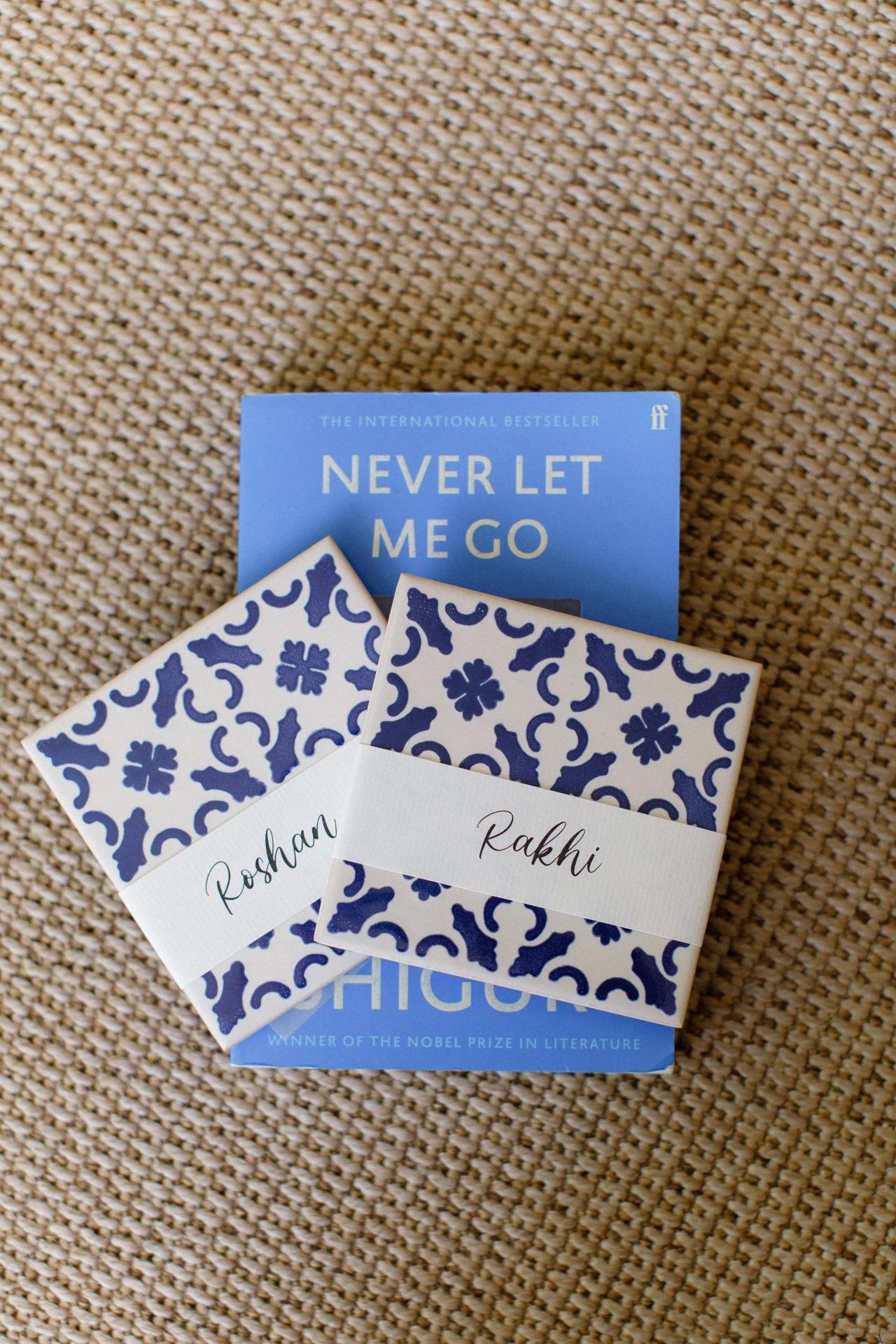 Blue book NEVER LET ME GO with two patterned ceramic coasters labeled Roshan and Rakhi on a textured fabric background.