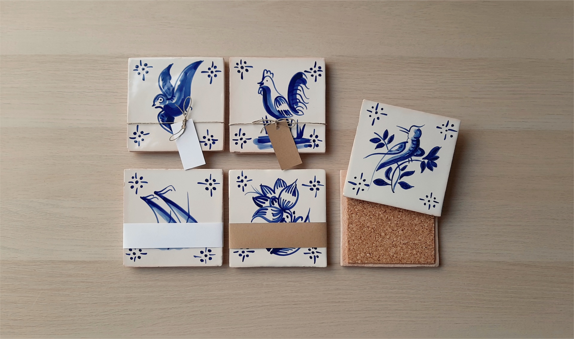 Ceramic coasters with blue hand-painted bird, rooster, flower, and grass designs, tied with paper bands and showing cork backing.