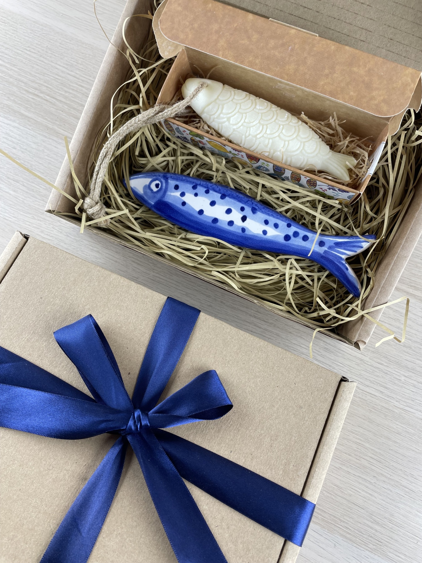 Two fish-shaped objects in a cardboard box with straw and a blue ribbon on a wooden surface