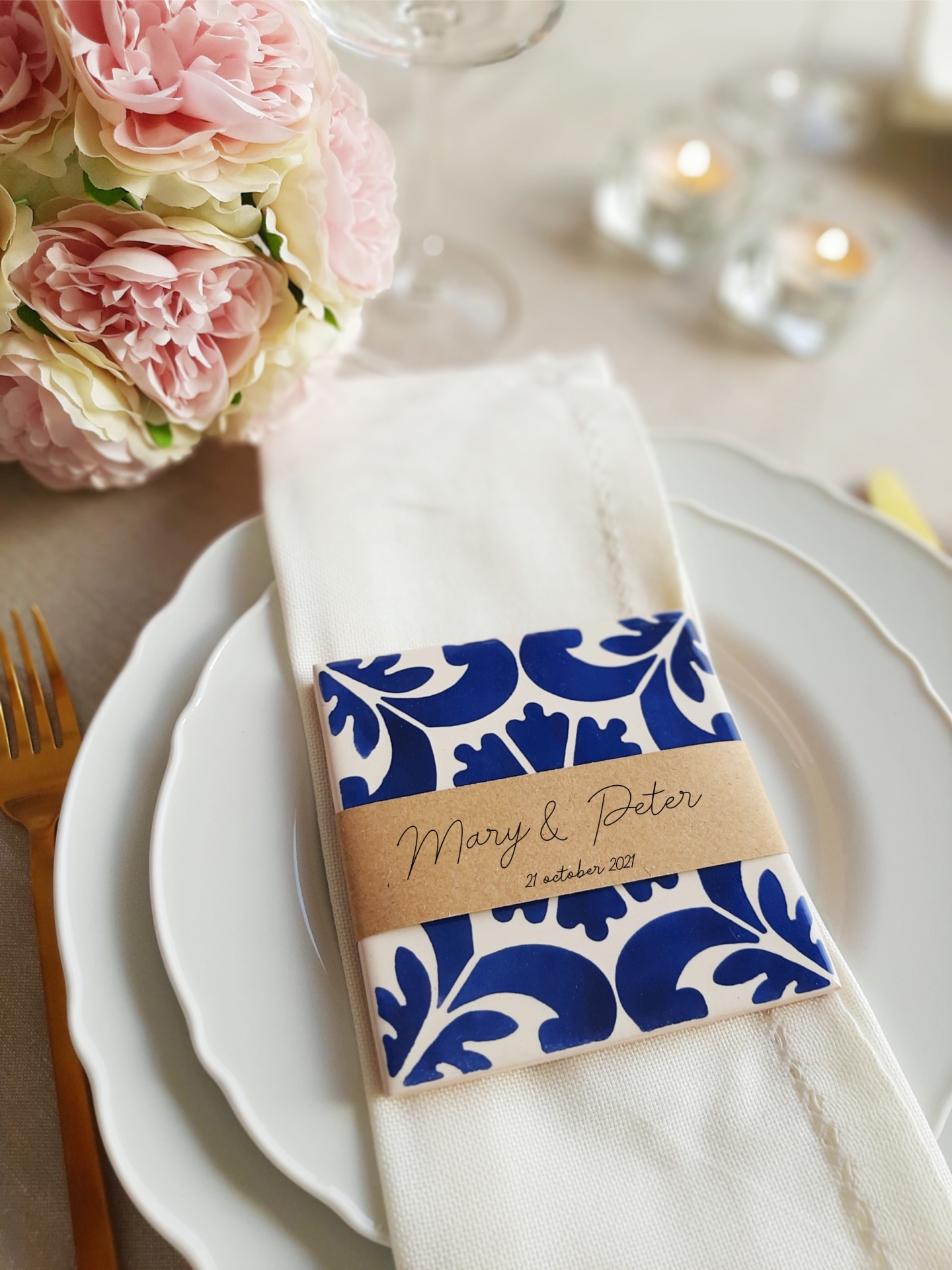 White napkin on white plates with blue patterned holder and brown sleeve saying Mary & Peter 21 october 2021, gold fork, pink roses
