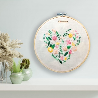 Bastidor de madeira com bordado floral em forma de coração pendurado numa parede cinzenta acima de uma prateleira branca com vasos e plantas