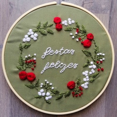 Bordado artesanal com flores vermelhas e brancas em tecido verde, com texto 'festas felizes' e aro de madeira