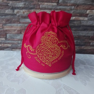 Saco vermelho de tecido com bordado amarelo e cordões, colocado sobre base branca com fundo muro de pedras.