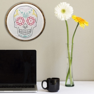 Secretária branca com portátil, caneca preta, vaso com flores e quadro de caveira bordado.